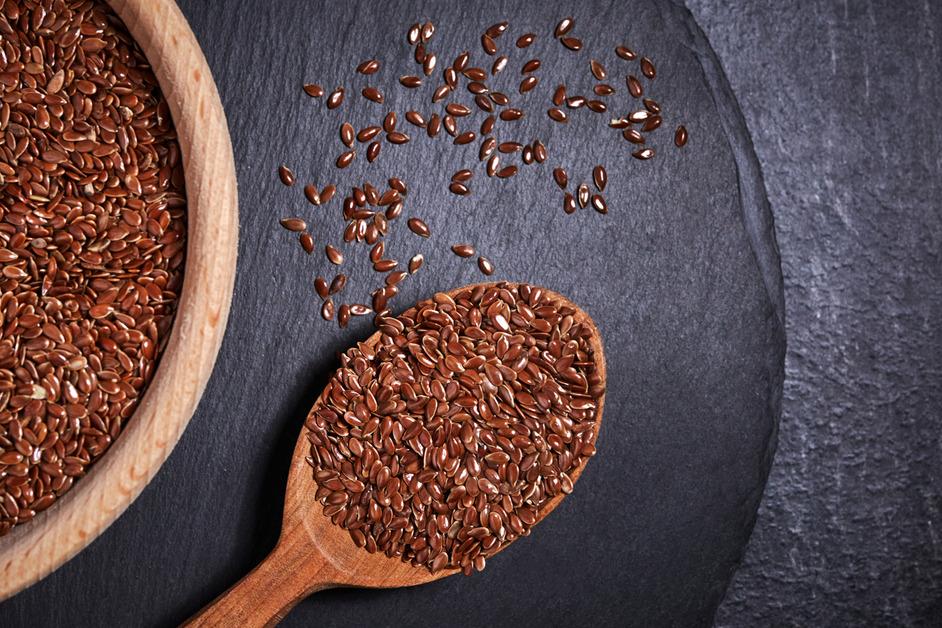 A spoonful of flax seed sits on a slate surface next to a a larger wooden bowl of flax seed that has been cut out of the frame.  