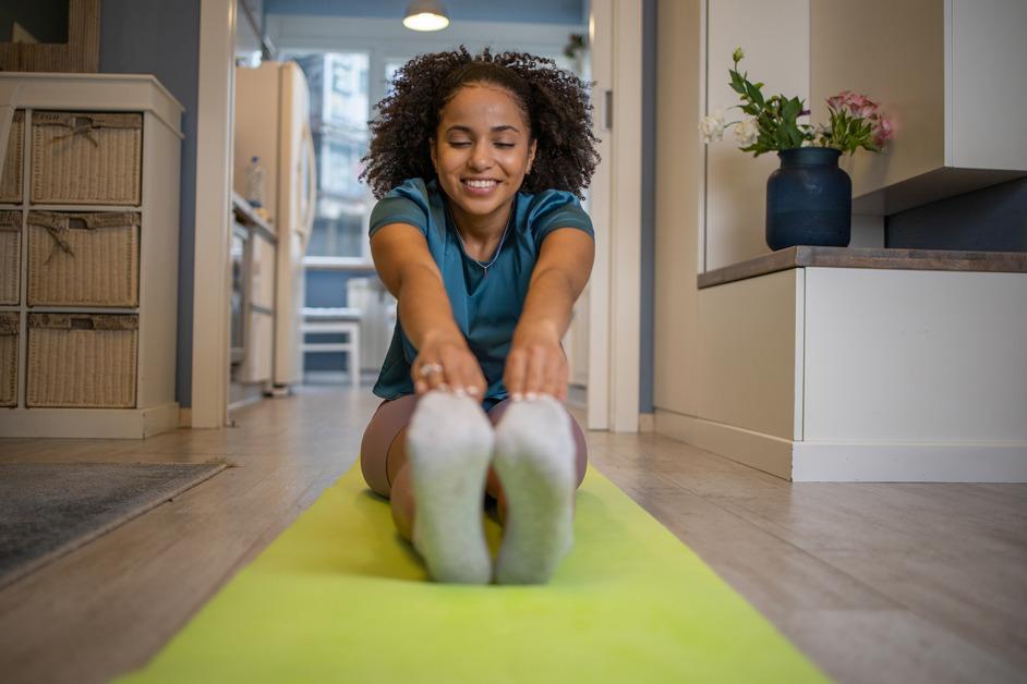 Woman sits on a yellow yoga mat and stretches both of her legs straight in front of her while smiling. 