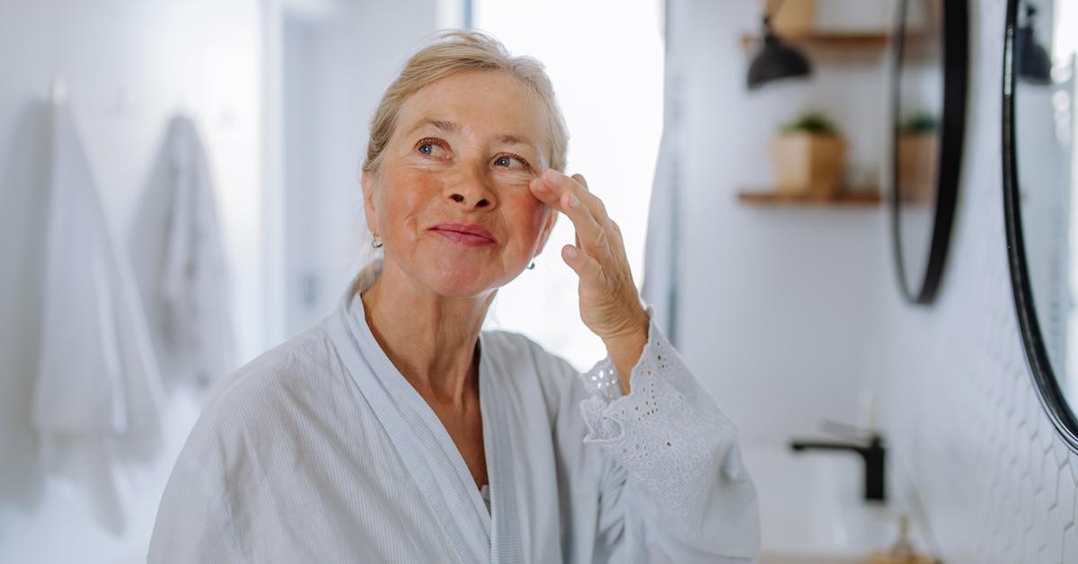 A senior woman wearing a white bathrobe dabs eye cream on her under eye area.