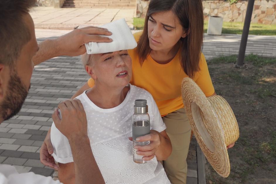 Senior woman experiencing heatstroke rests against younger woman while man presses a cold towel to her forehead. 