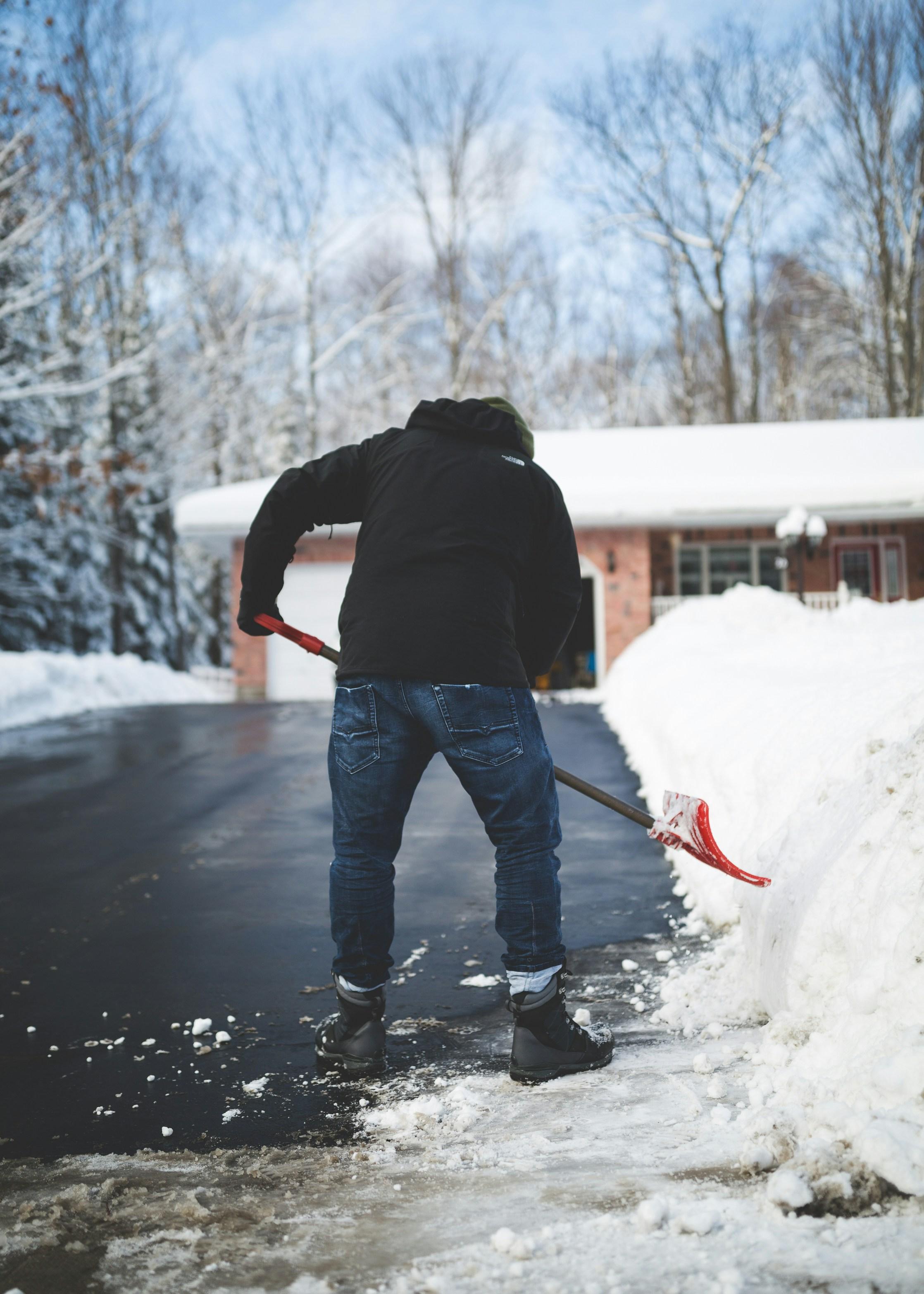 A photo depicts a man from the neck to his knees wearing a black jacket and jeans while shoveling snow with a red shovel in front of a house.