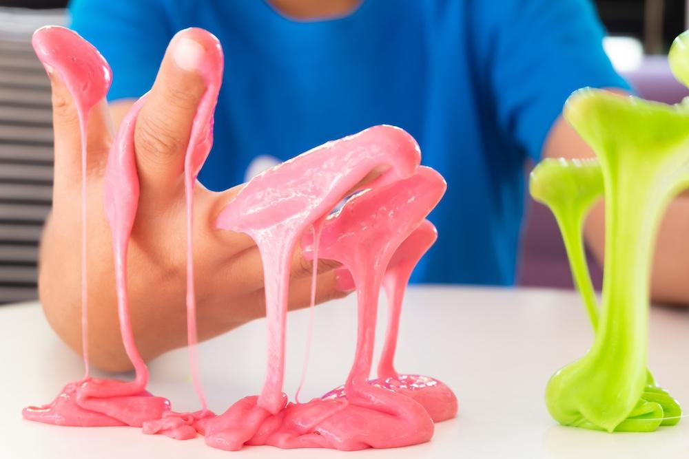 A kid wearing a blue shirt shows hands covered in pink and green slime.