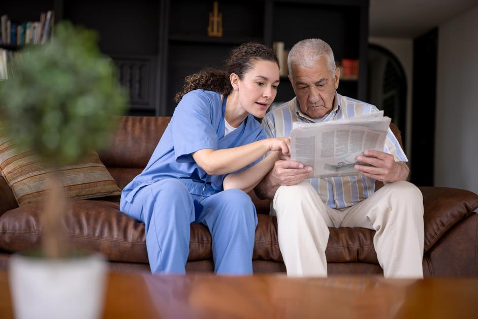 A nurse wearing blue scrubs sits on a brown leather couch next to a senior man and points to the newspaper he is reading. 