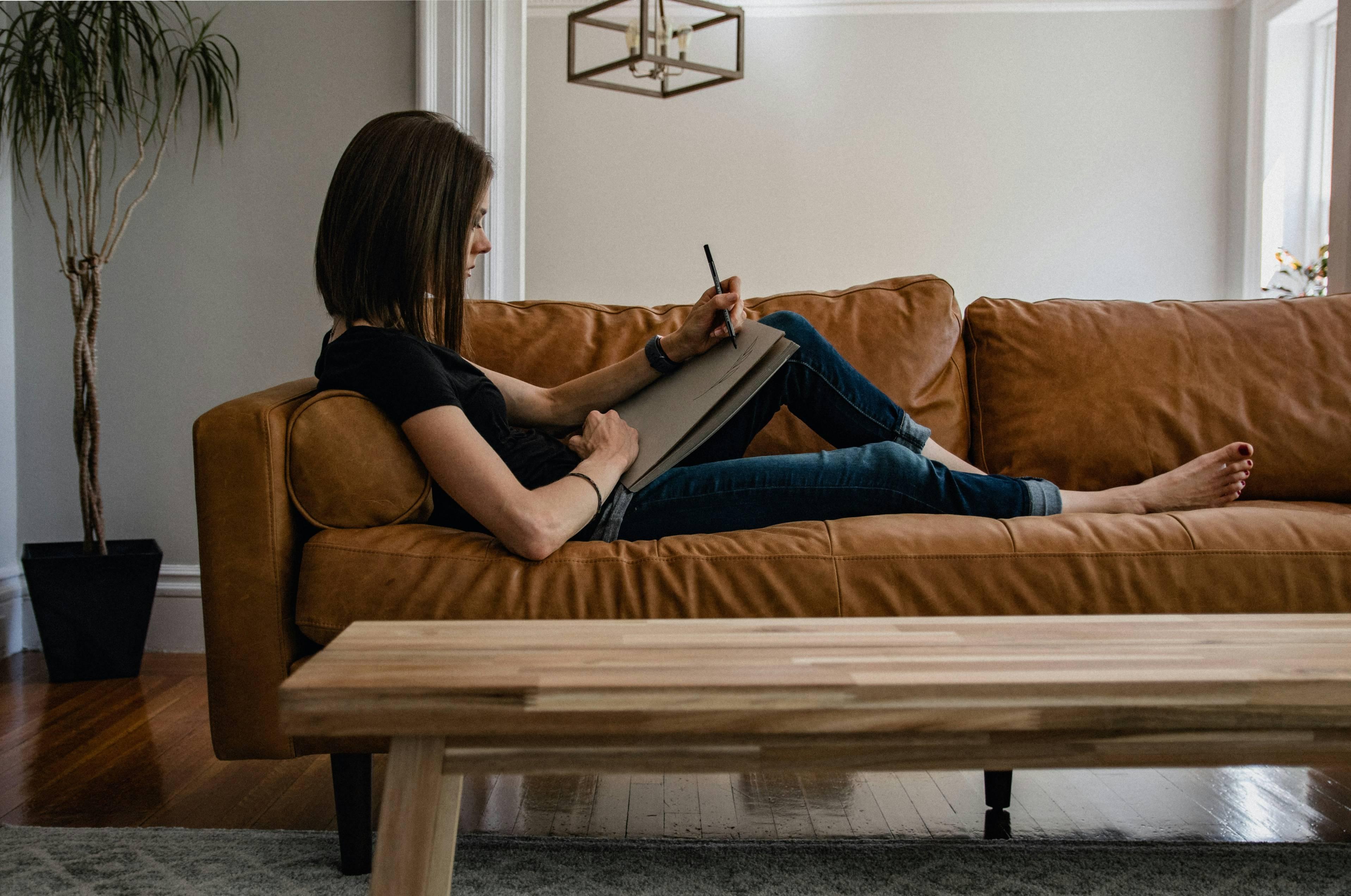 A woman draws on a piece of paper while sitting barefoot on her couch.