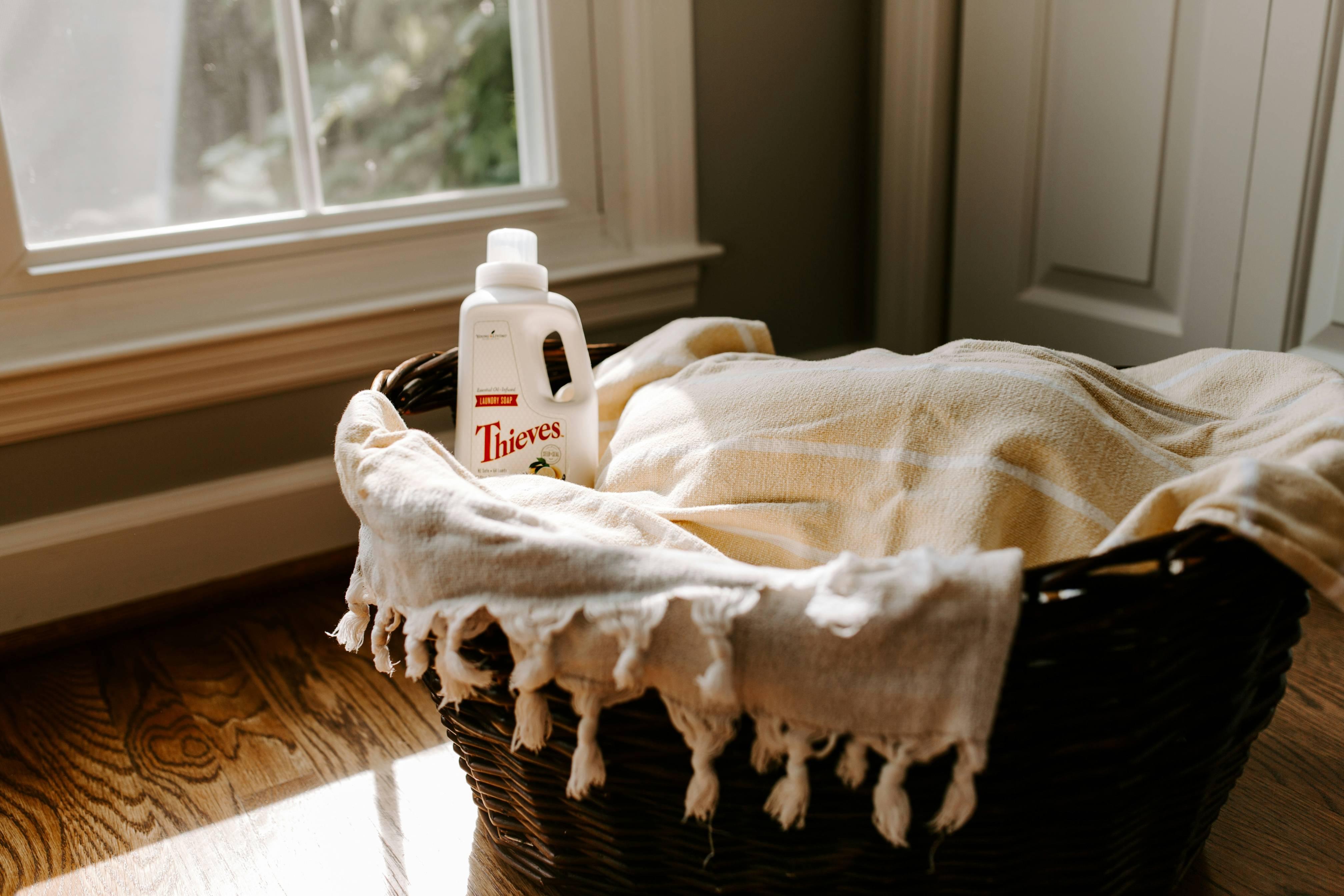 A wicker laundry basket atop a wooden floor is pictured with a throw blanket inside of it along with a bottle of laundry detergent.