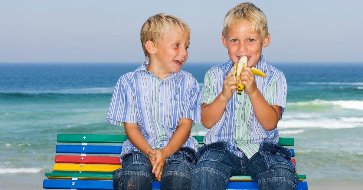 Two boys sitting on a bench by the ocean with one eating a banana. 