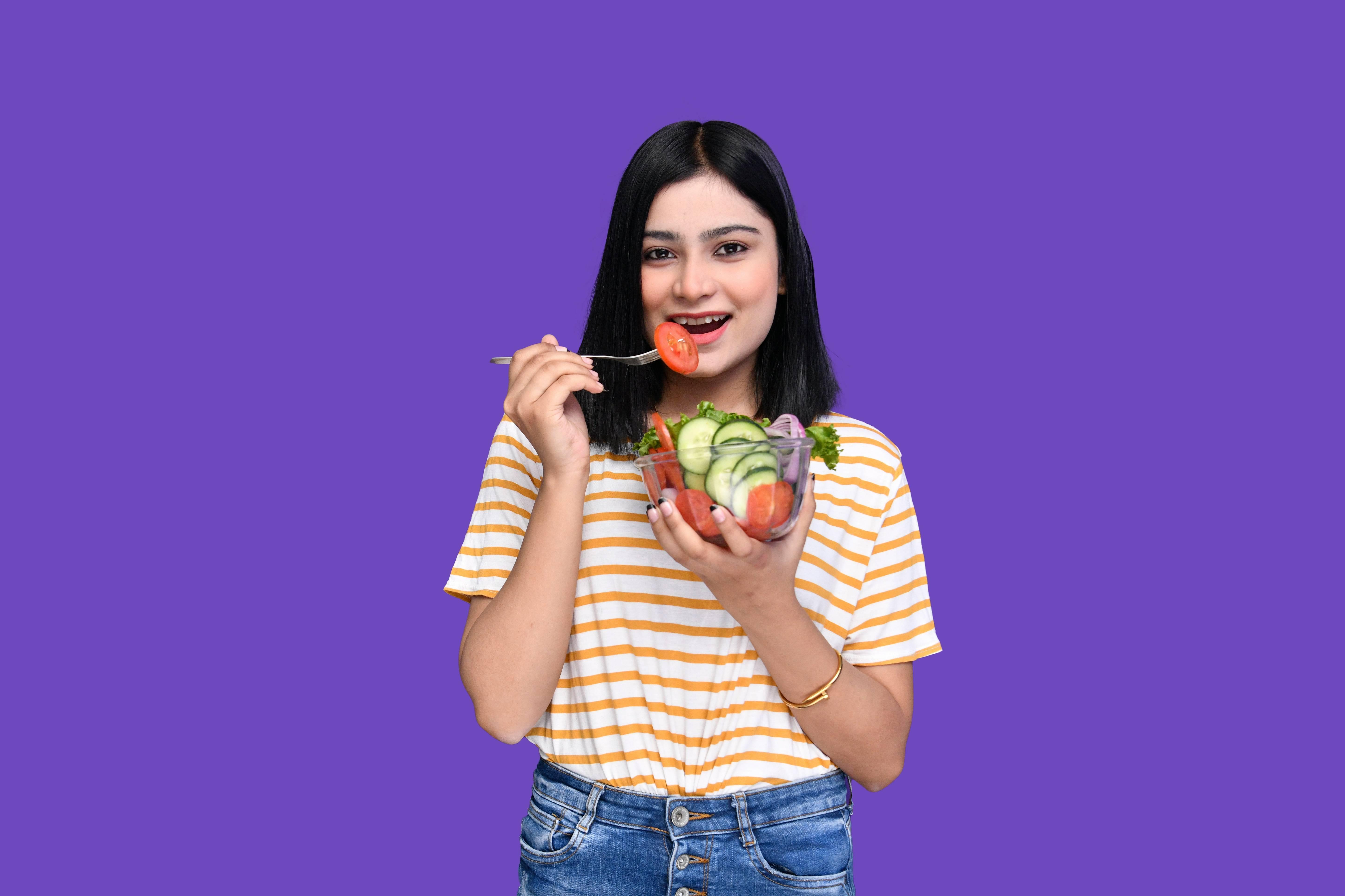 A smiling woman holds a bowl of vegetables as she prepares to eat one with a fork.