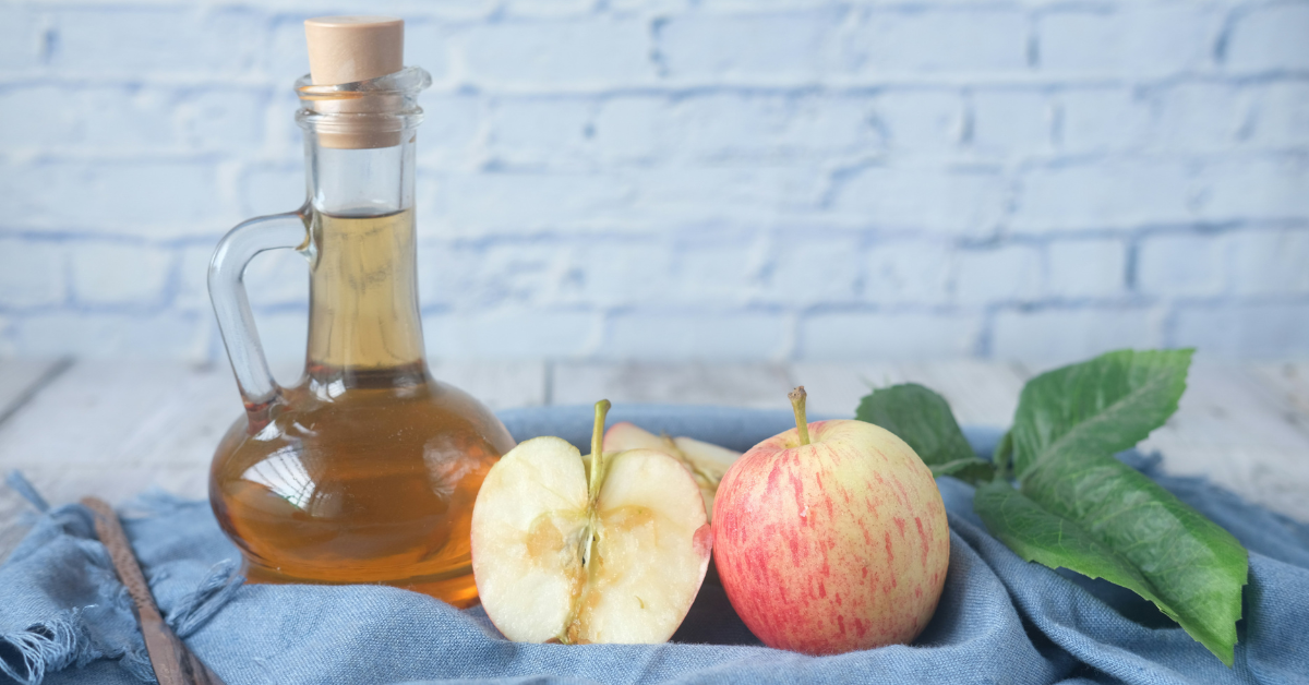 A decanter of apple cider vinegar sits on a blue cloth with some apples