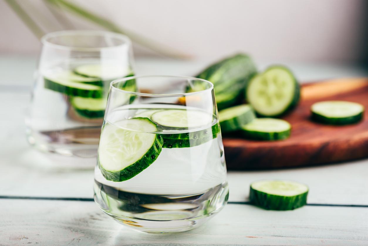 Two glasses of cucumber water atop a table next to a cutting board with cucumbers on it.