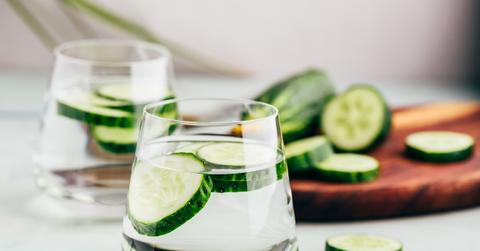 Two glasses of cucumber water atop a table next to a cutting board with cucumbers on it.