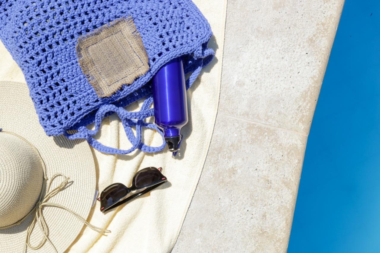 A pair of sunglasses, a sun hat, a water bottle, and a purple crocheted beach bag all laid out on a towel near a pool.