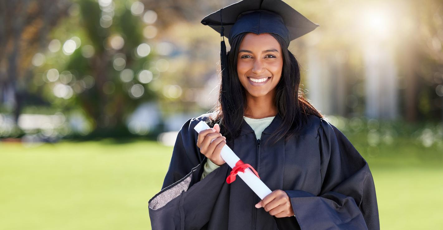 A smiling woman in a black gown poses with her master's degree diploma at her school.