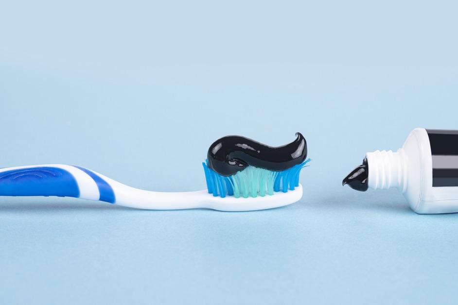 A close up photo of a toothbrush in line with a bottle of black toothpaste in front of a blue background.  