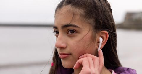 Young girl with brown hair and purple jacket stands at the beach while touching her AirPod in her ear.