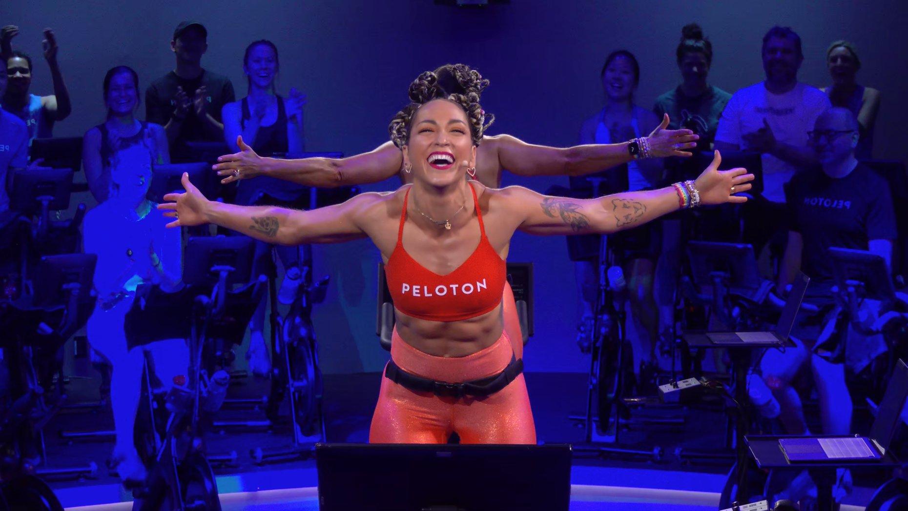 A Peloton instructor smiles while teaching from atop a Peloton bicycle with members behind her.