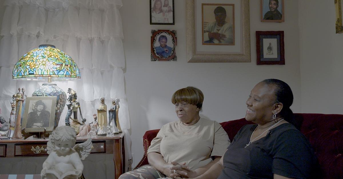 Still from Resita Cox's documentary 'Freedom Hill' showcasing two women sitting on a burgundy couch in front of a wall adorned with photographs