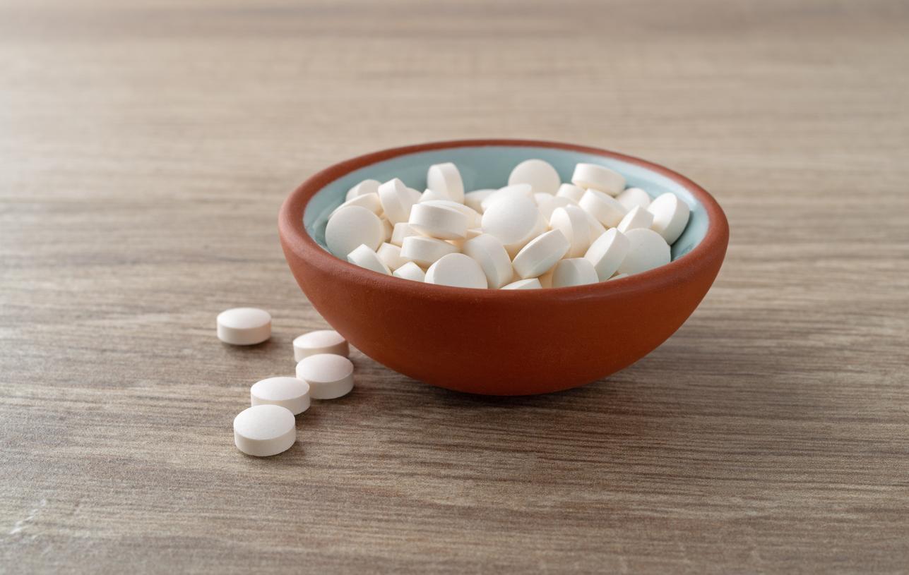 A red and blue bowl contains white DHEA pills with five pills spilled beside the bowl on a wooden table.