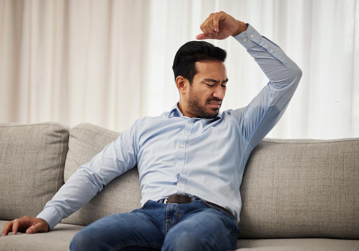 A man sits on the couch and makes an unpleasant facial expression while sniffing his underarms from his wet button-up shirt.