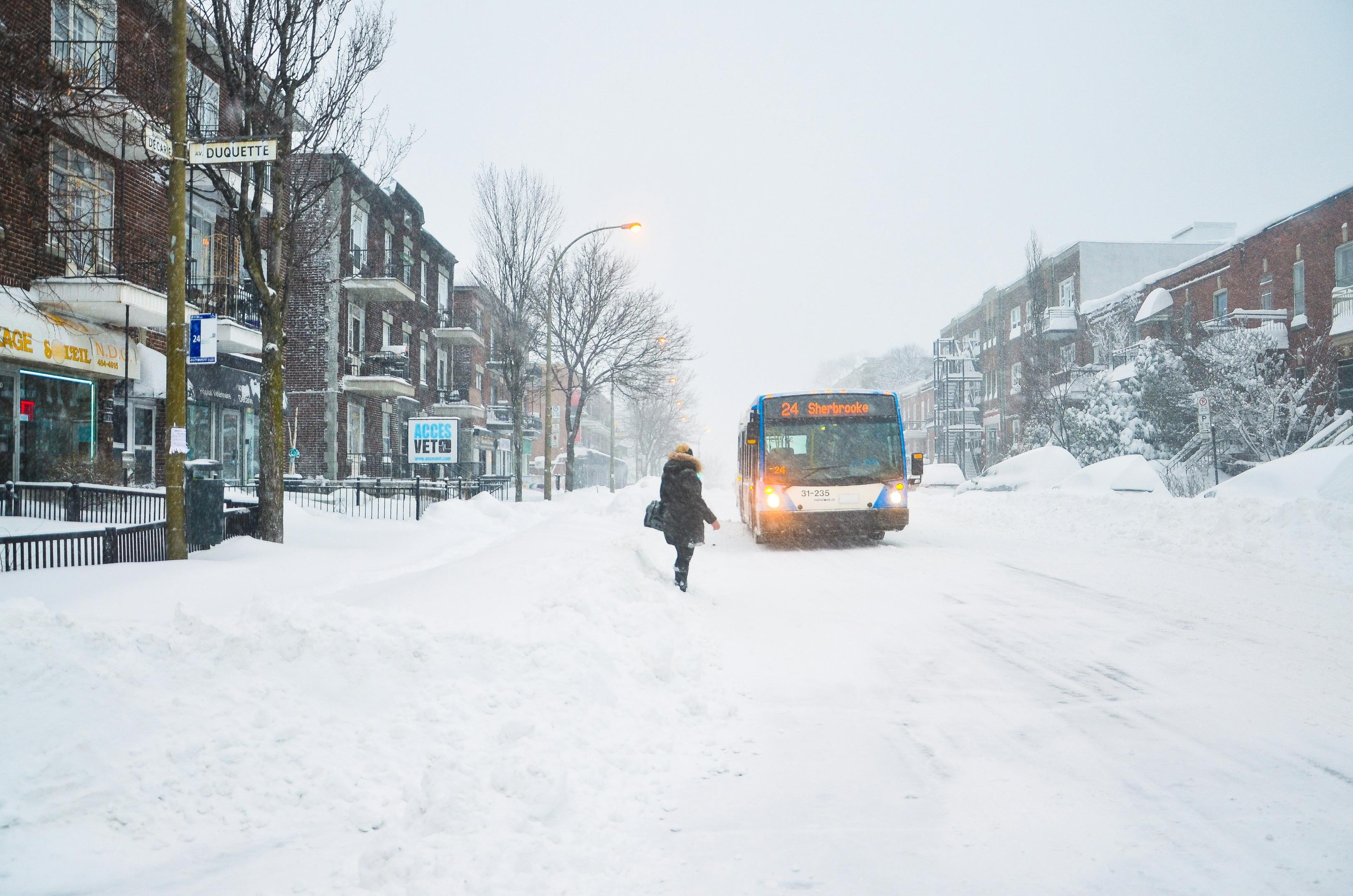 A woman wearing a heavy winter coat waits for an oncoming bus driving on a snowy road inside a village where it has snowed heavily.