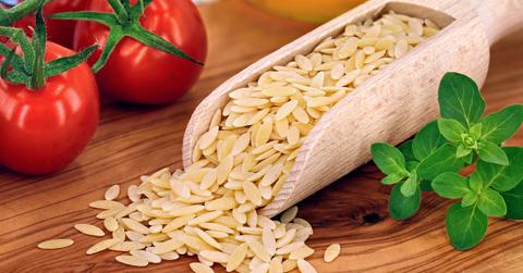 Orzo pasta spilling over from a wooden spoon onto a table next to tomatoes.