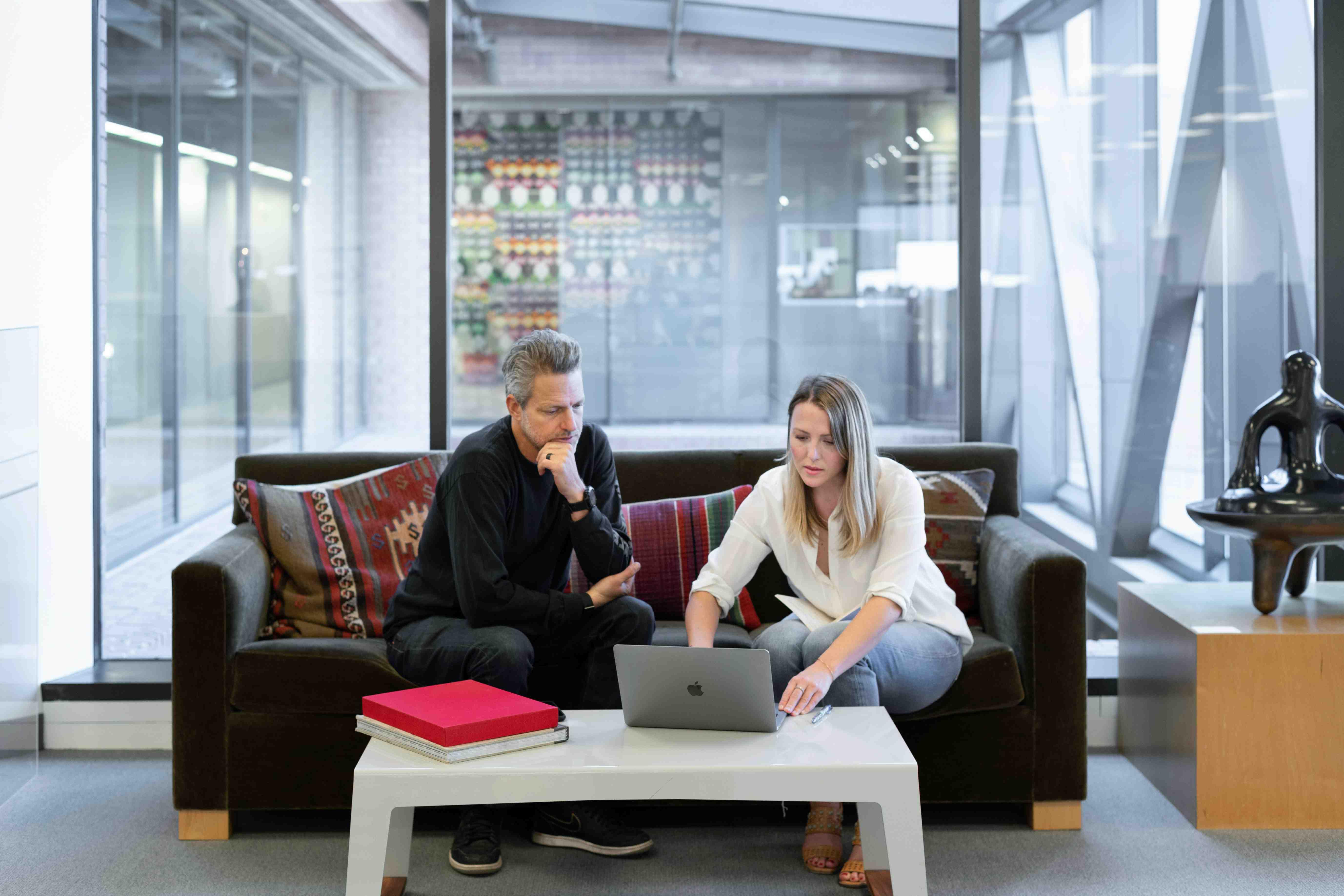 Two employees sit together in an office, working on a laptop.