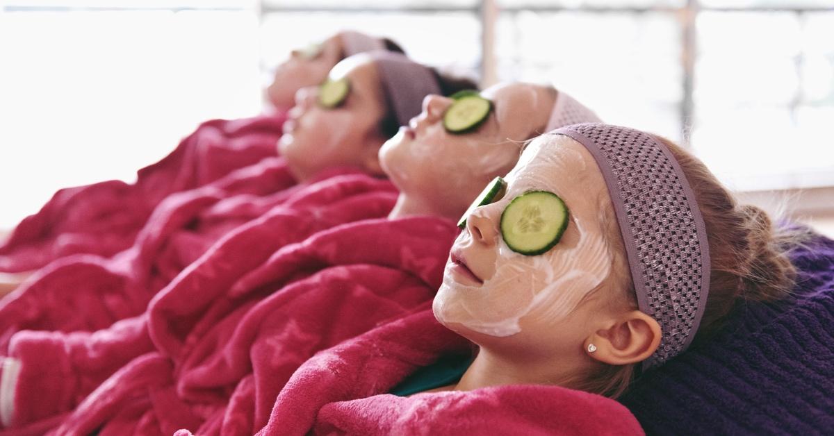 Four girls sitting in a row wearing magenta robes, face masks, and cucumbers on their eyes.