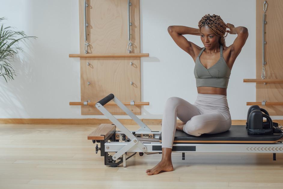 A woman in a gray workout outfit sits on the edge of a reformer while tying her hair up. 