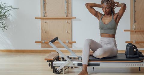 A woman in a gray workout outfit sits on the edge of a reformer while tying her hair up.