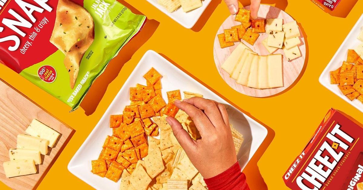 Photo of two hands reaching for Cheez-It crackers and slices of cheese on plates and wooden boards.