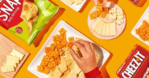 Photo of two hands reaching for Cheez-It crackers and slices of cheese on plates and wooden boards.