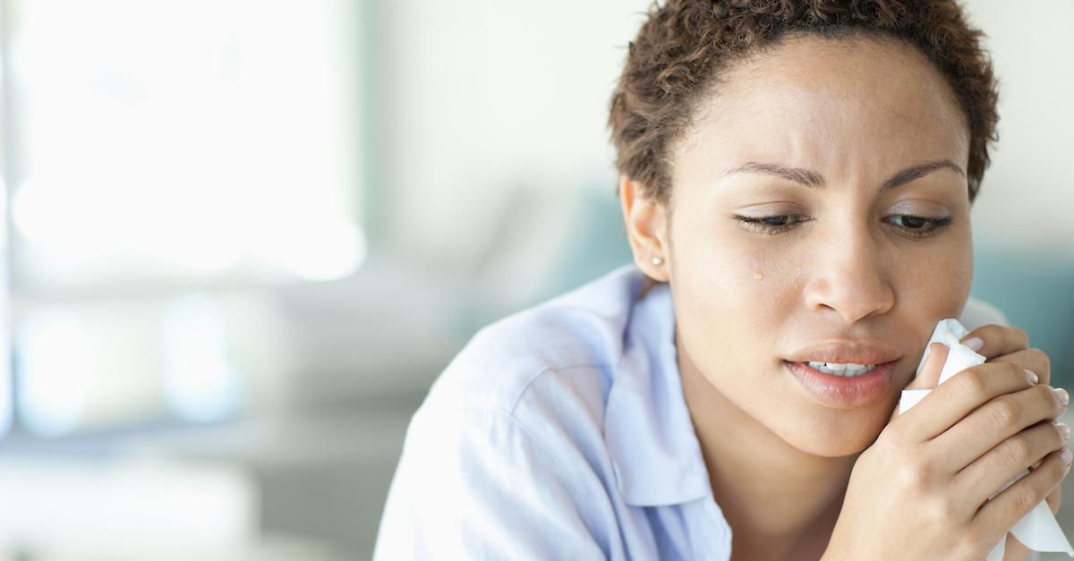 A person with short hair in a light blue button-down is crying, and holds a tissue to her face