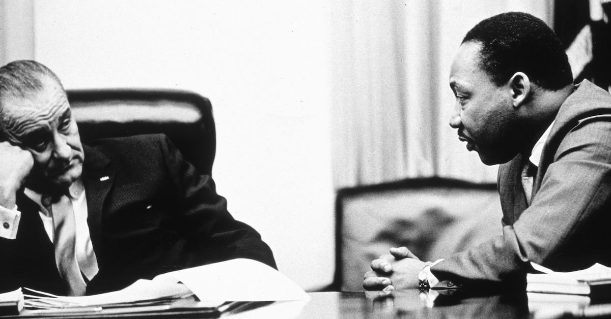 Martin Luther King Jr. and Lyndon B. Johnson speak at a desk, in a black-and-white image.