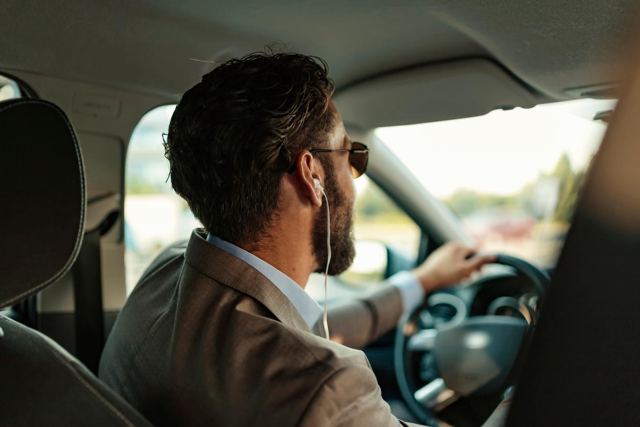 A man wearing white earphones is photographed from behind while driving a car.