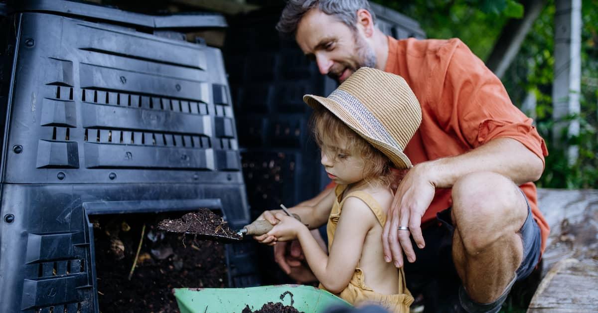 A parent with their child, shovelling compost into a small wheelbarrow.