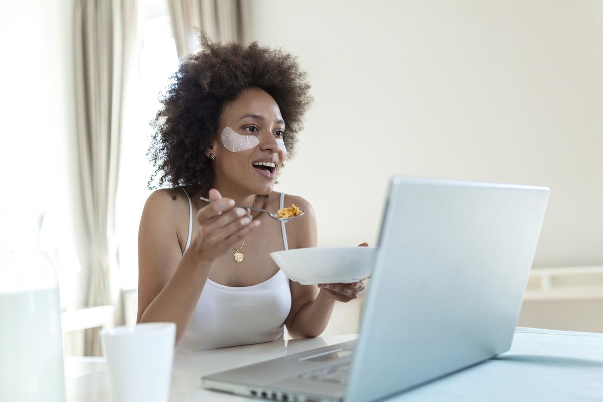 Woman eating cereal with eye patches and looking at her laptop 