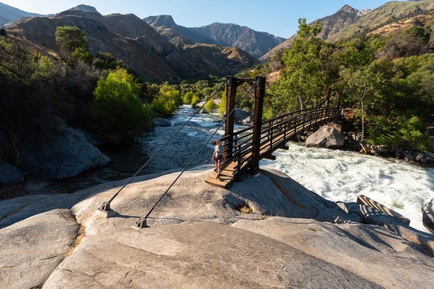 A woman in summer hiking clothes stands at the wooden hanging bridge at Potwisha Campground in Sequoia National Park surrounded by rocks,  trees, and mountains.