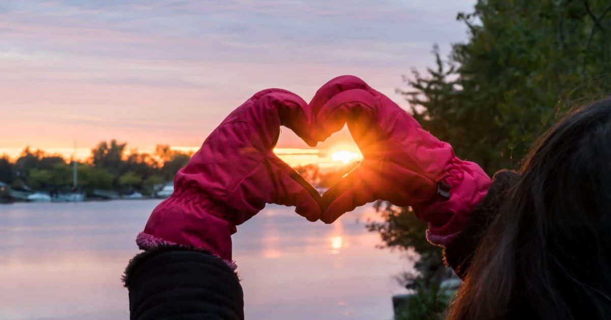 Sunset beams of light shine through heart-shaped hands of a woman looking at winter landscape. (Representative Cover Image Source: Getty Images | Milaspage) 