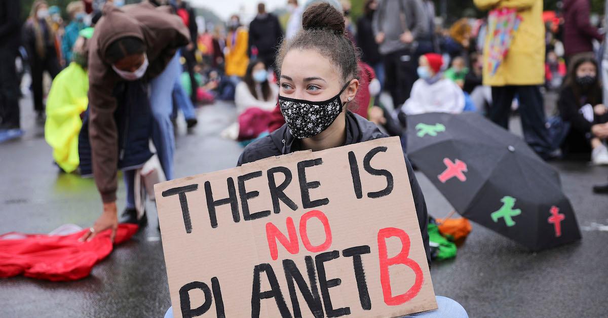 A protester sits on the ground wearing a mask, holding a sign that reads "there is no planet B."