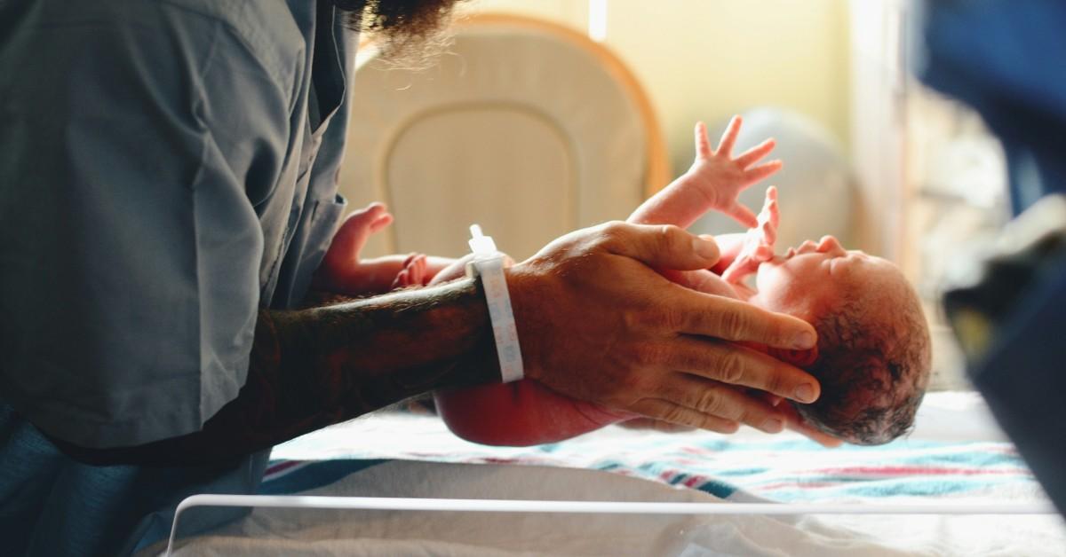 A new dad holds his baby over a bassinet while wearing a hospital bracelet 