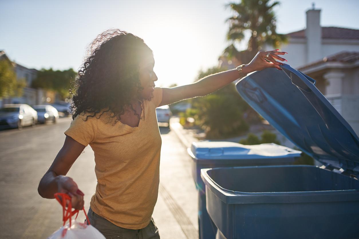 A woman opens her blue trash bin while holding a trash bag in her other hand.