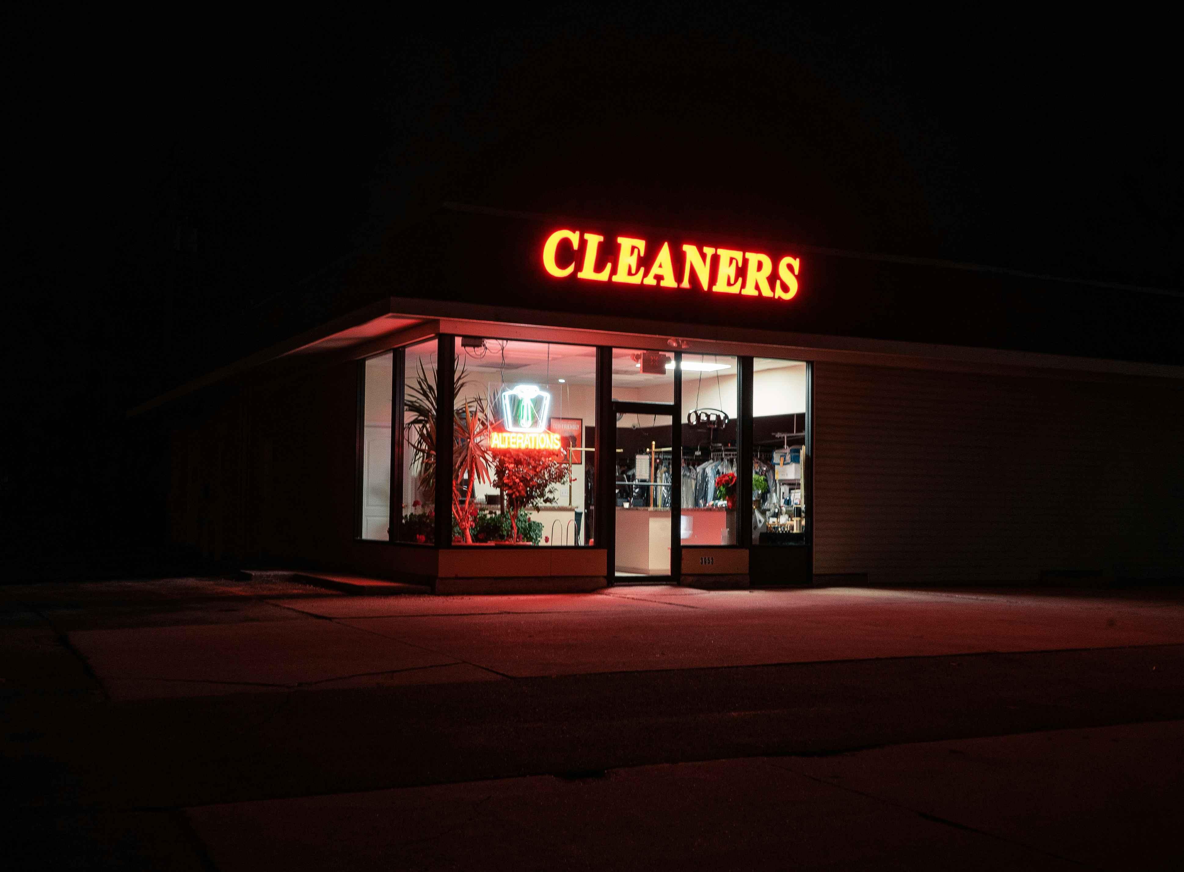 A dry cleaning store is pictured at night illuminated by a red-orange light that reads "Cleaners".