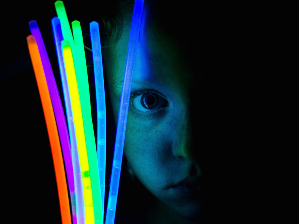 A close-up portrait photo of a child holding seven glow sticks of various colors.