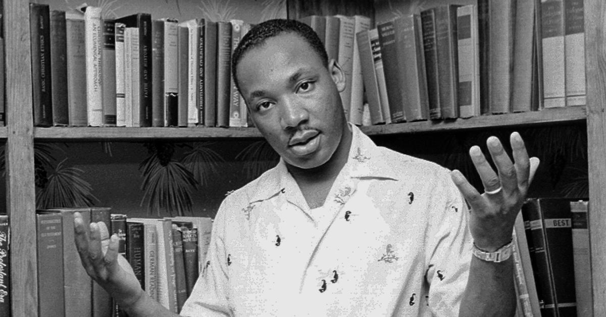 Martin Luther King Jr. poses with his hands up in front of a bookcase in a black-and-white image.