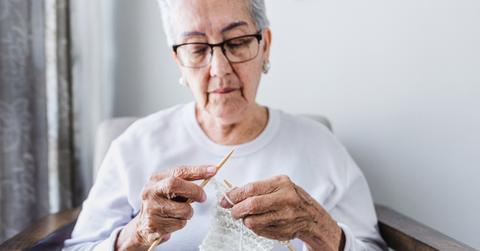 Person holding two knitting needles and working on a project made of white yarn while sitting down.