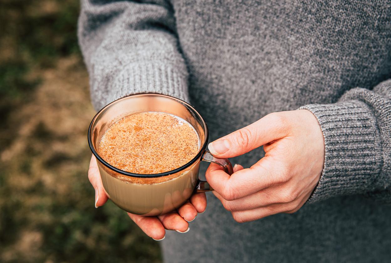 A person in a gray sweater holds an amber cup of chai tea seasoned with cinnamon.