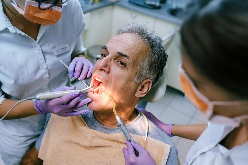 A man sits in a chair at the dentist with his mouth open while two dentists use tools in his mouth. 