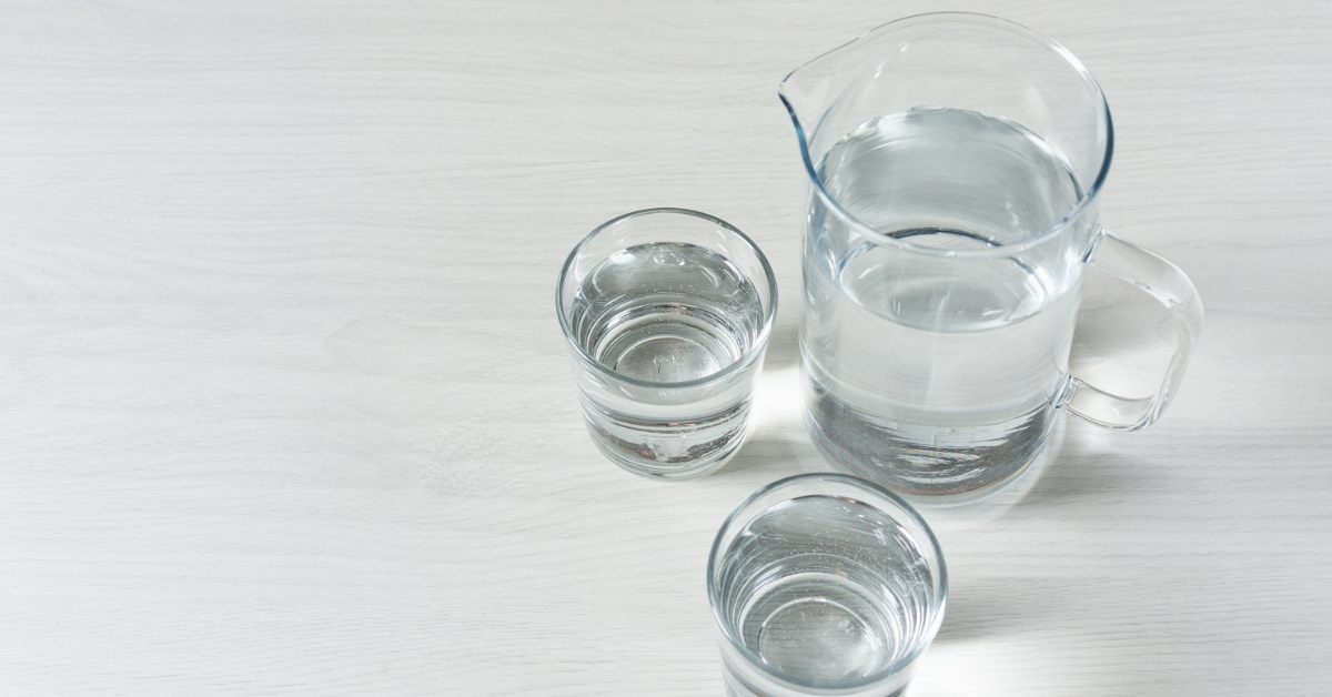 A pitcher with two glasses of water sit on a gray background 