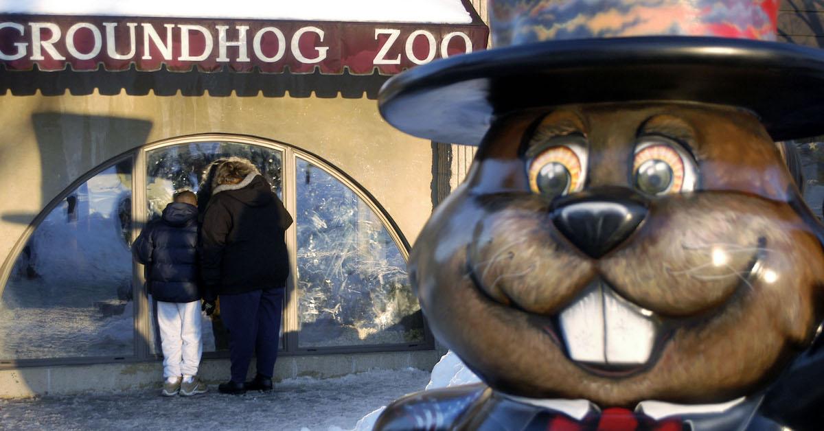 Two people look into Punxsutawney Phil's enclosure at Groundhog Zoo, with a wooden groundhog statue in the foreground