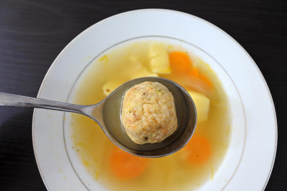 A spoon holds a matzah ball over a bowl of soup