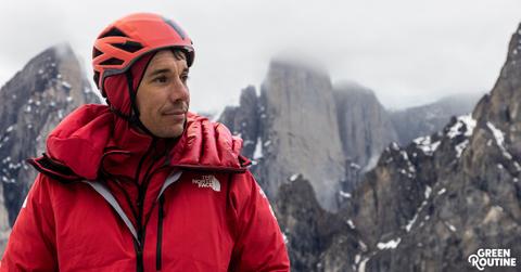 Alex Honnold wears an orange helmet and red jacket with snowy mountains in the distance